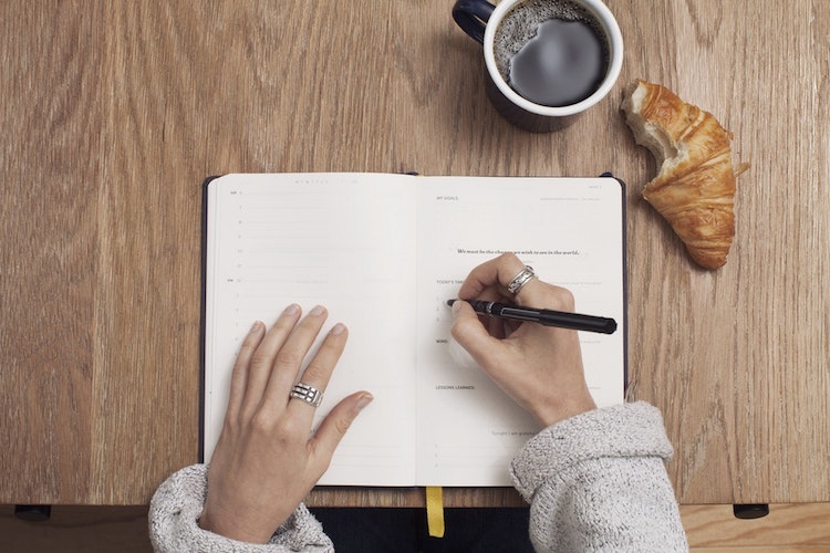 A person writing in a journal, eating a croissant, and drinking a cup of coffee. 